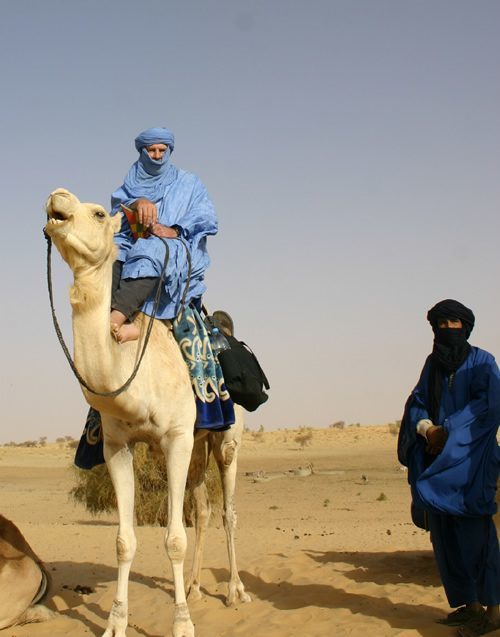 Mounted on camel in Sahara with a Tuareg dressed in blue standing nearby.