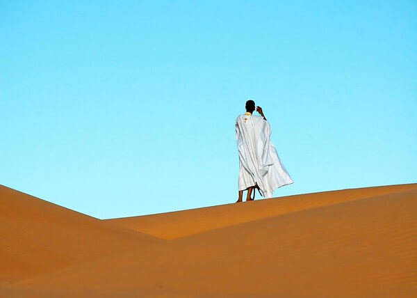 Man in a white robe standing on a sand dune looking off to the distance.