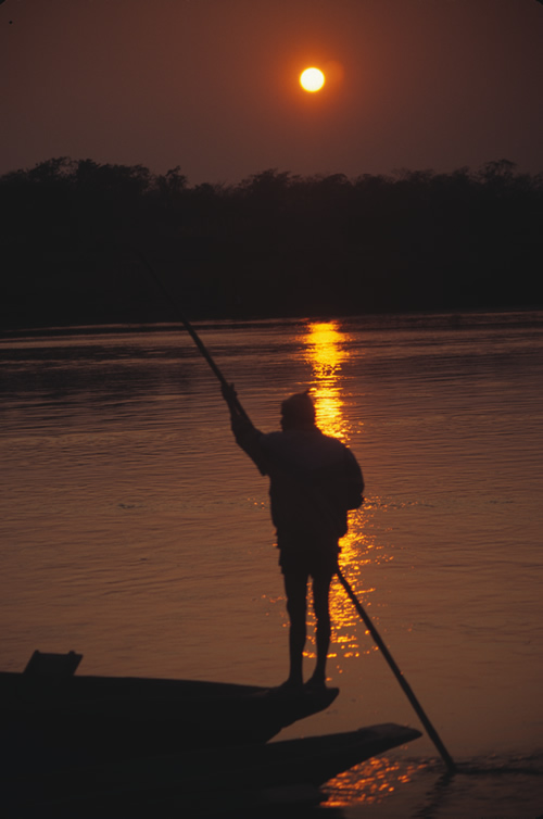 A standing man rowing a canoe on a river in Nepal at sunset.